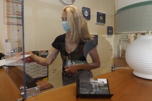 Un área de recepción con una recepcionista detrás de una mampara de vidrio, un reloj en la pared y un jarrón blanco grande sobre una mesa.