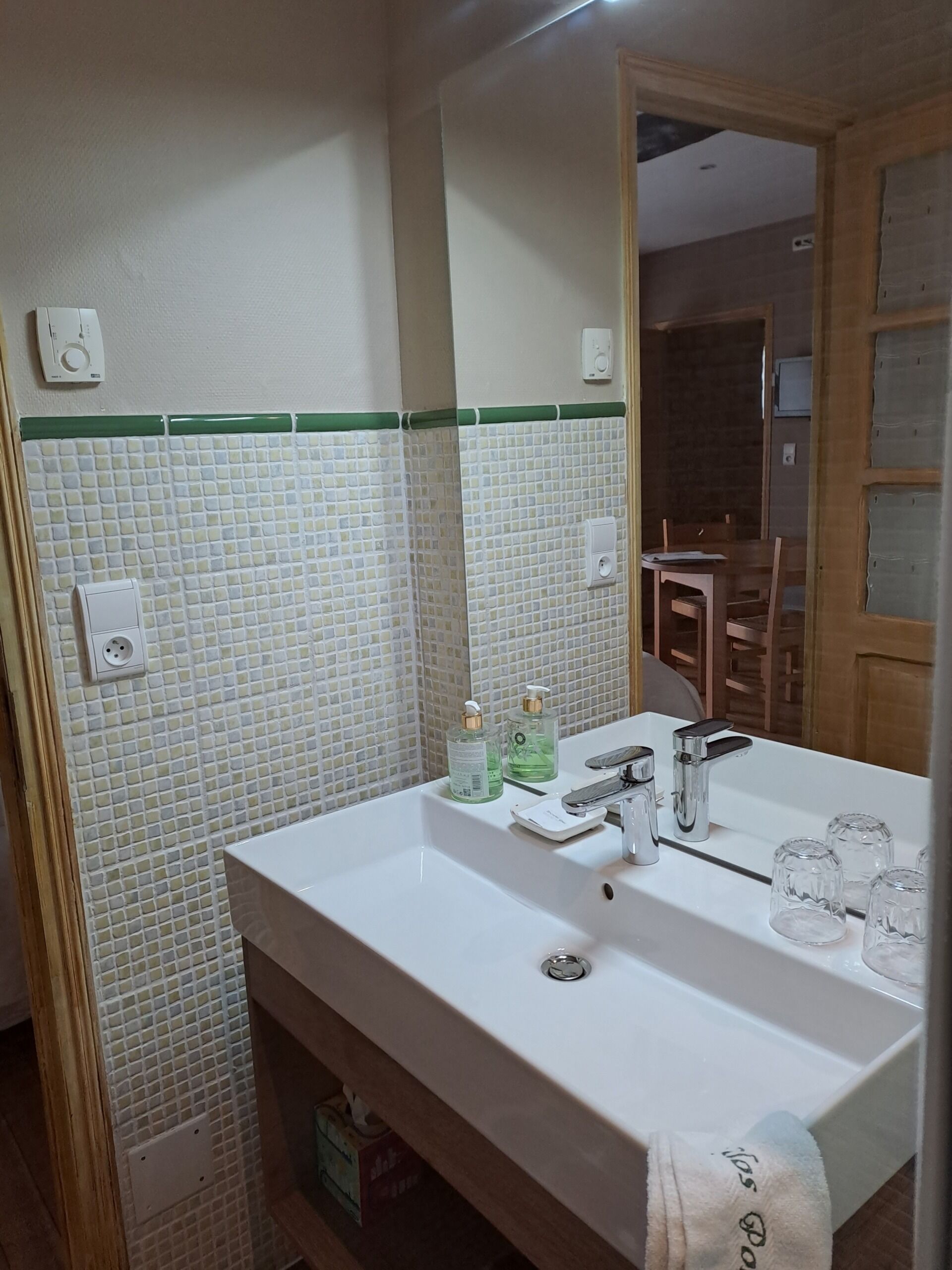 A bathroom with a white sink, green-tiled backsplash, and a mirror. There are two bottles of liquid soap and a towel hanging on the wall.