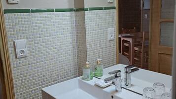 A bathroom with a white sink, green-tiled backsplash, and a mirror. There are two bottles of liquid soap and a towel hanging on the wall.