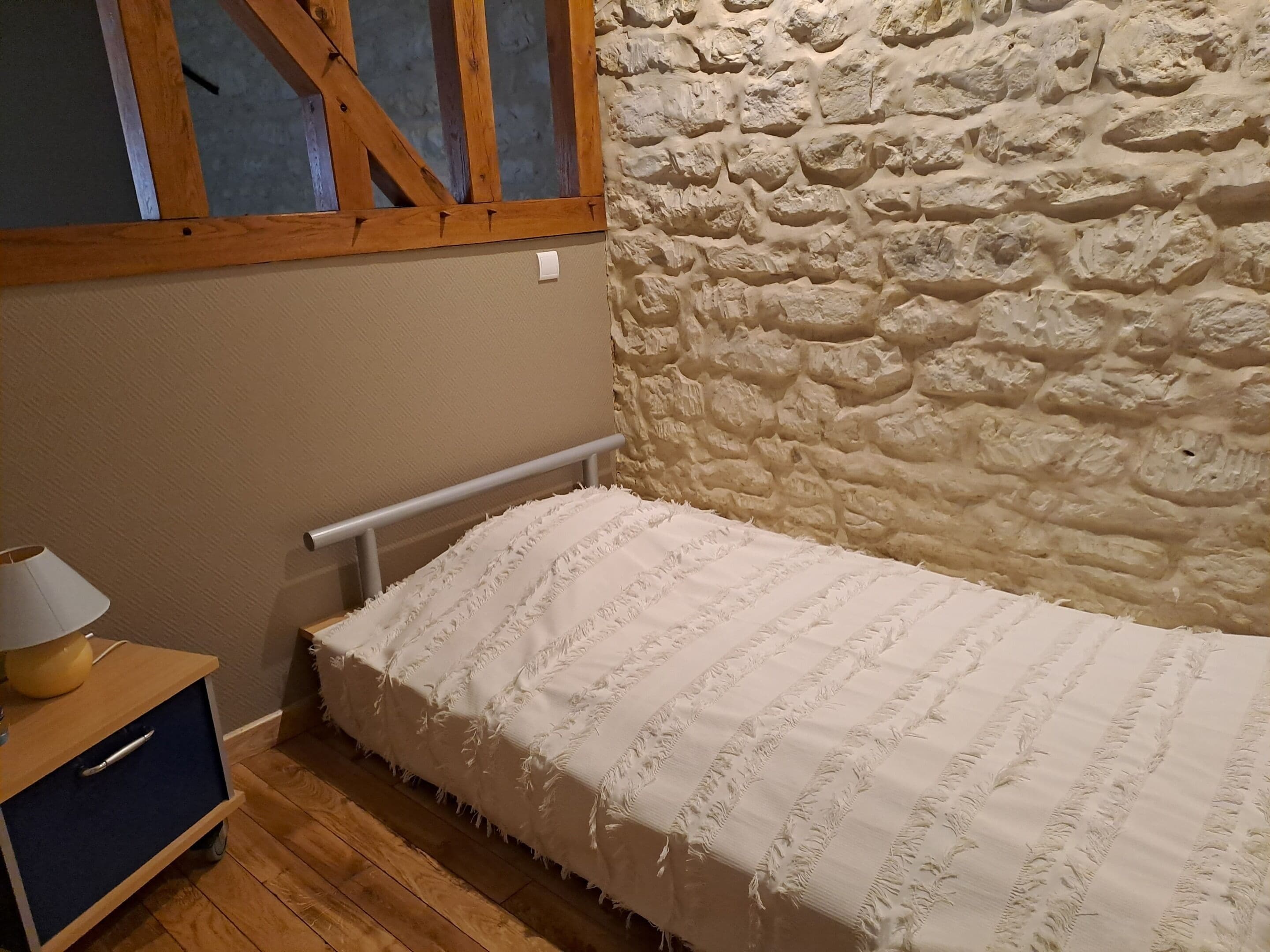 A small, cozy room with a stone wall, a wooden bed frame, and a nightstand with a lamp.