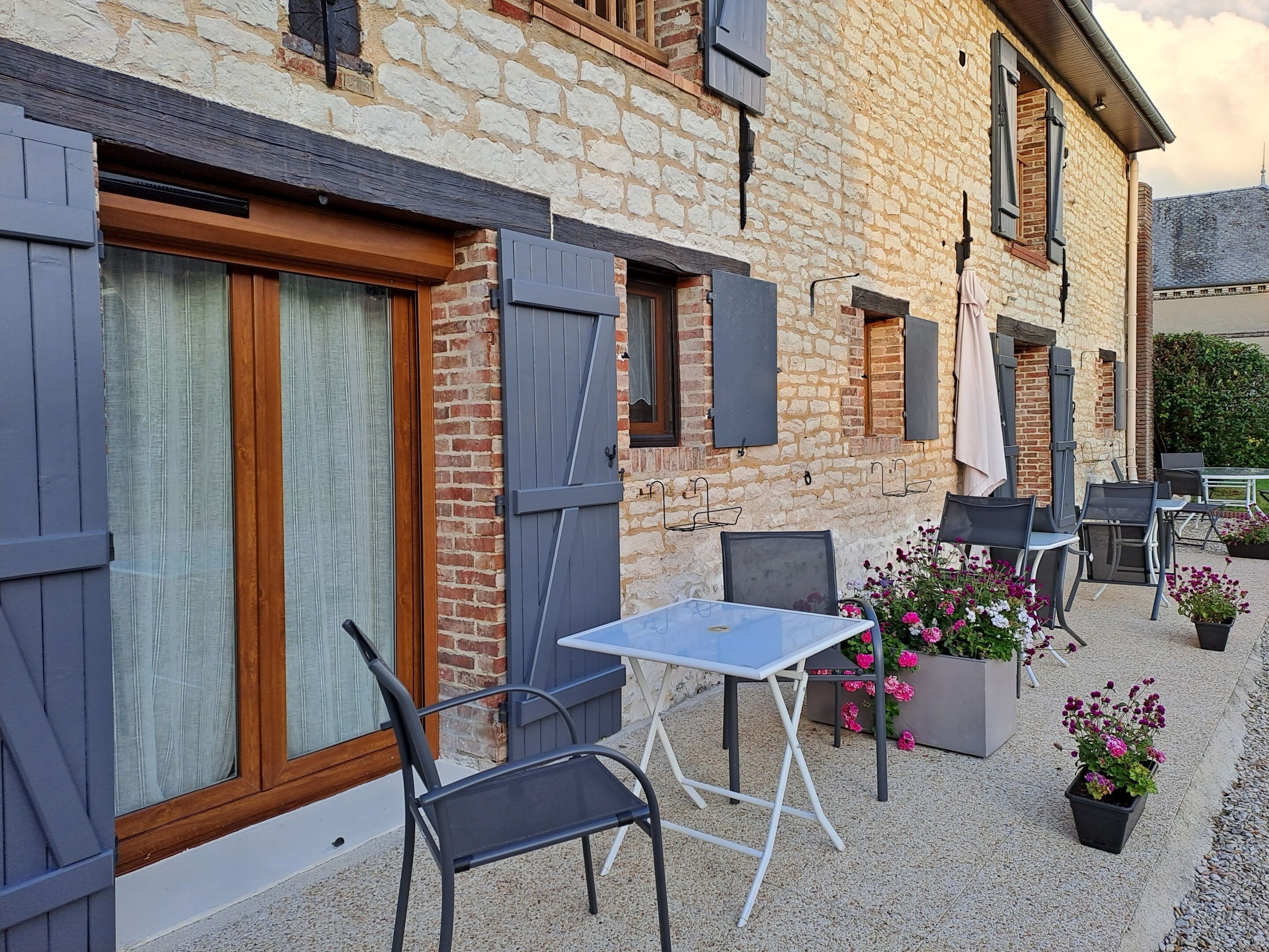 An outdoor patio with a table and chairs, a brick building with shutters, and potted flowers.