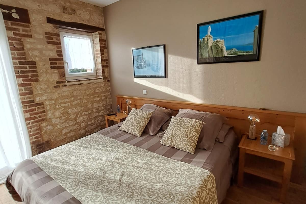 Ein Schlafzimmer mit Backsteinwand, einem Fenster, einem Bett mit gemusterten Bettwaren, zwei Nachttischen und zwei gerahmten Bildern an der Wand.