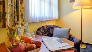 A hotel room with a dining table set with fruits, a newspaper, and a bottle of Gerolsteiner water. There is a chair with a patterned cushion and a lamp on the side. - Hotel Garni Fock Und Oben (Hamburg)