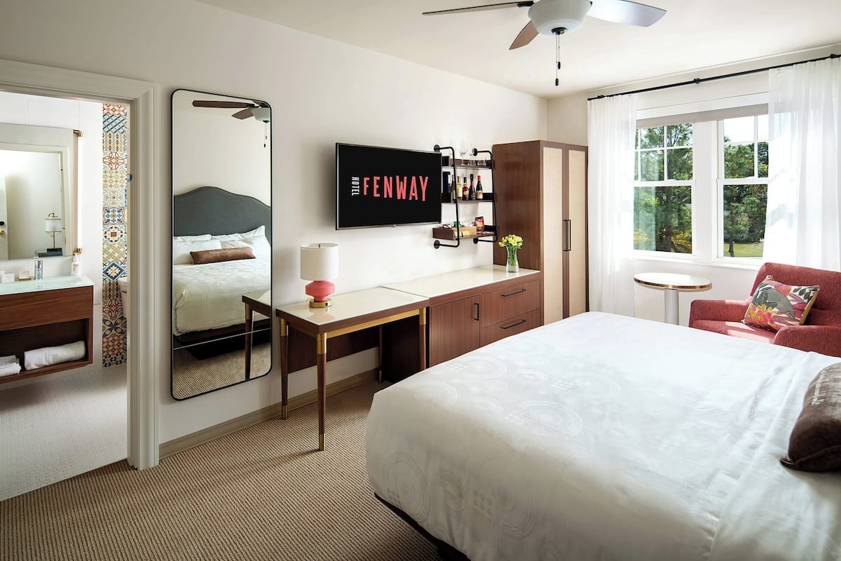A hotel room with a bed, a desk, a television, a wardrobe, a mirror, and a window with a view of trees.