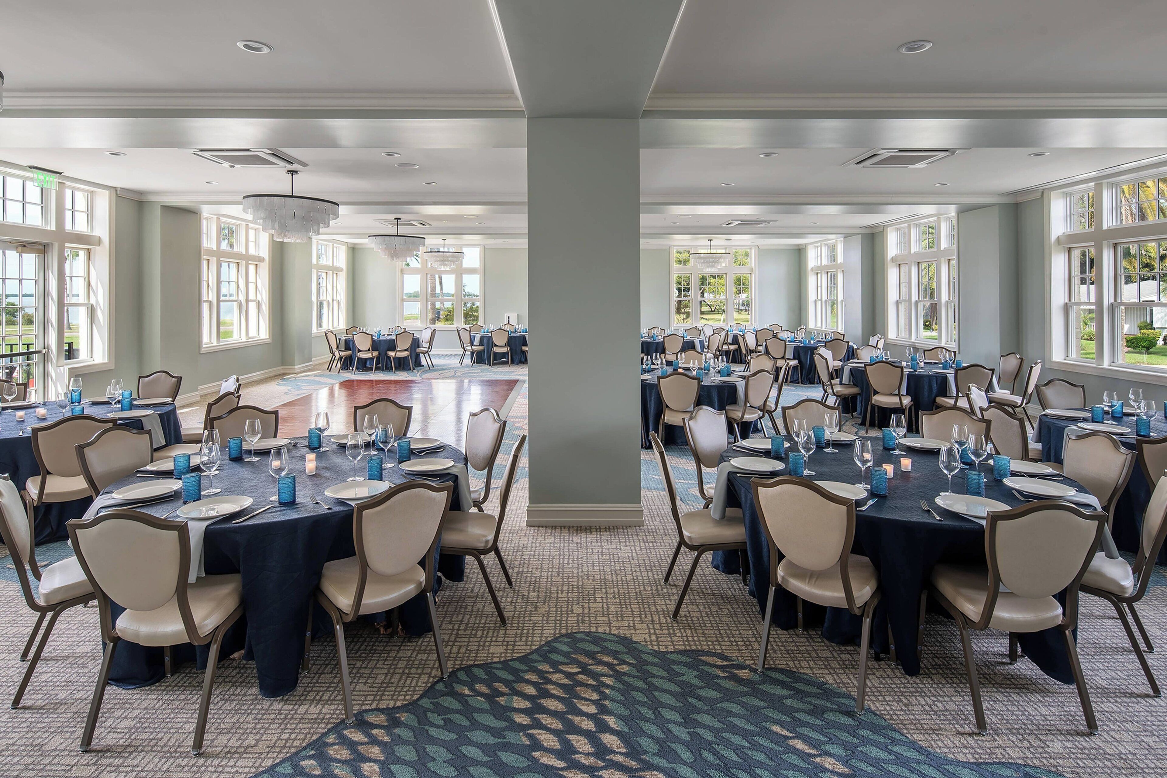 A large banquet hall with multiple round tables set for dining, featuring beige chairs and dark tablecloths.
