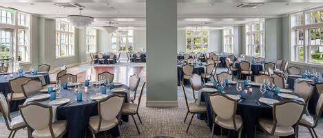 A large banquet hall with multiple round tables set for dining, featuring beige chairs and dark tablecloths.