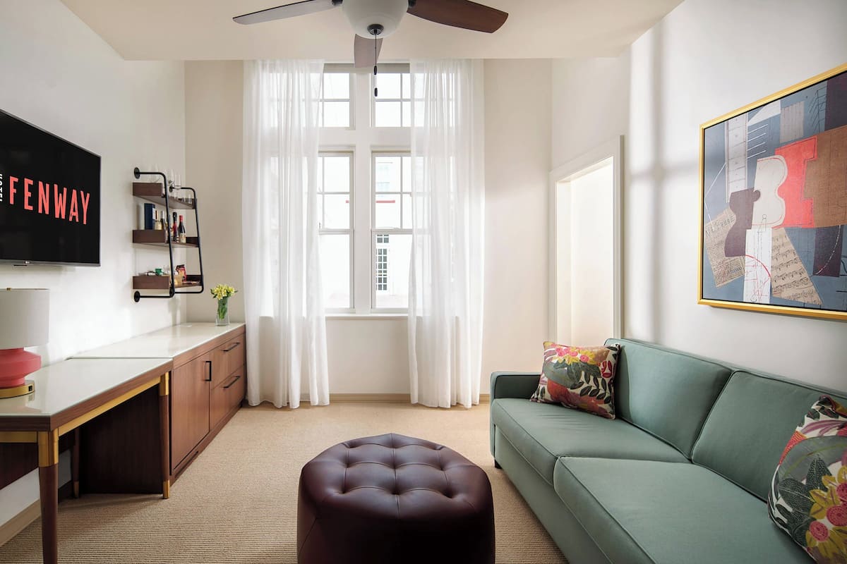 A modern living room with a flat-screen TV, a green sofa, a wooden desk, and a large window with sheer curtains.