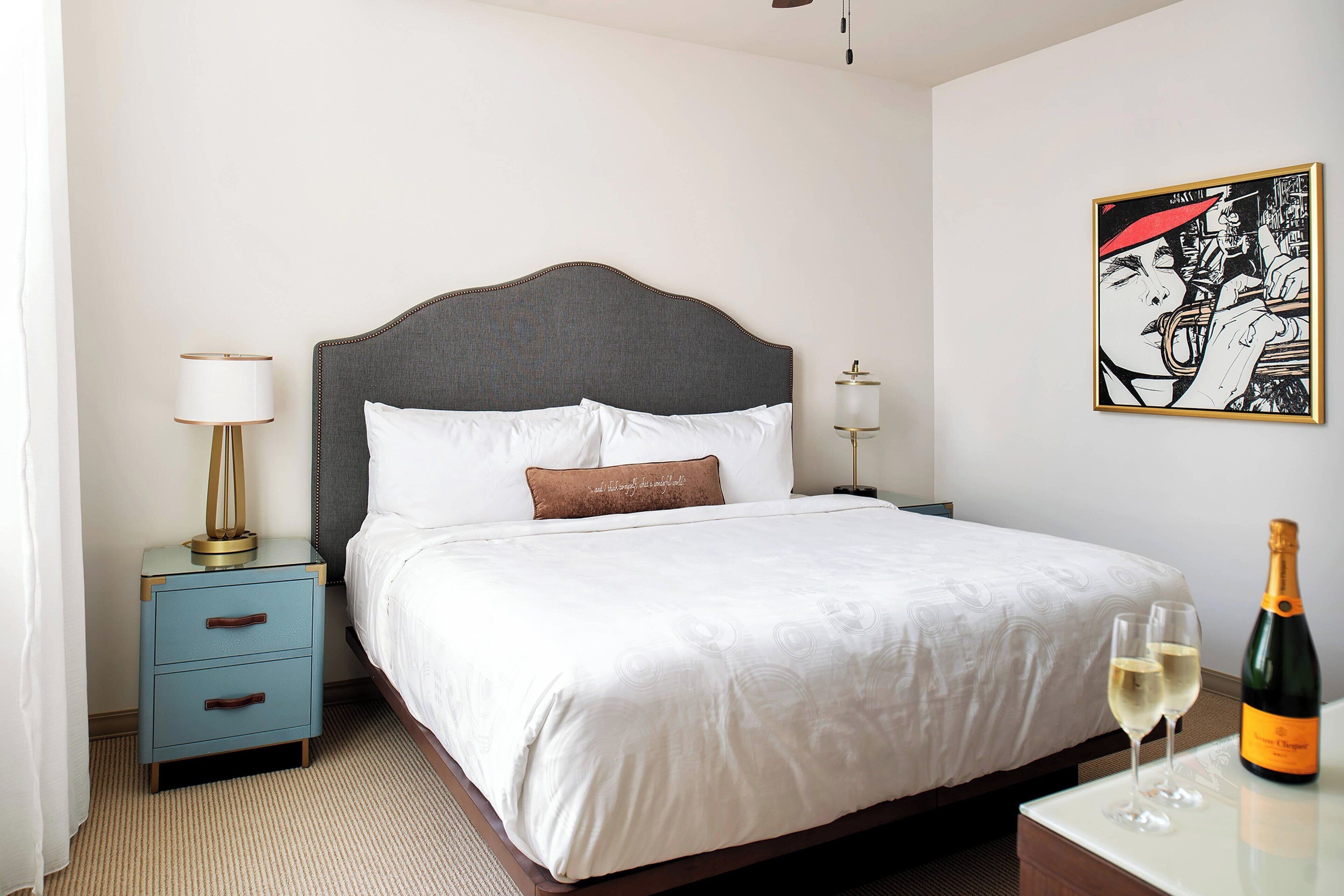 A neatly made bed with white linens, a nightstand with a lamp, and a bottle of champagne on a table.