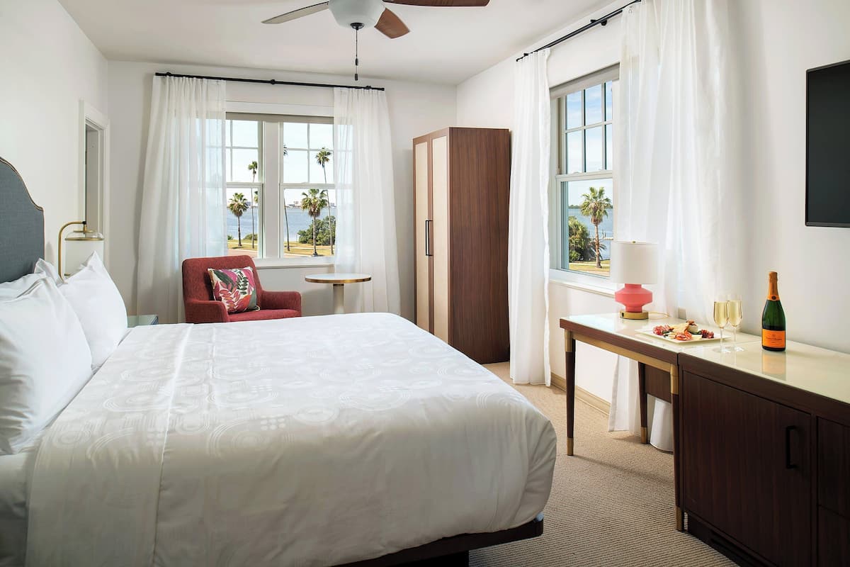 A hotel room with a large bed, a desk with a bottle and glasses, a red chair, and a view of palm trees through the windows.