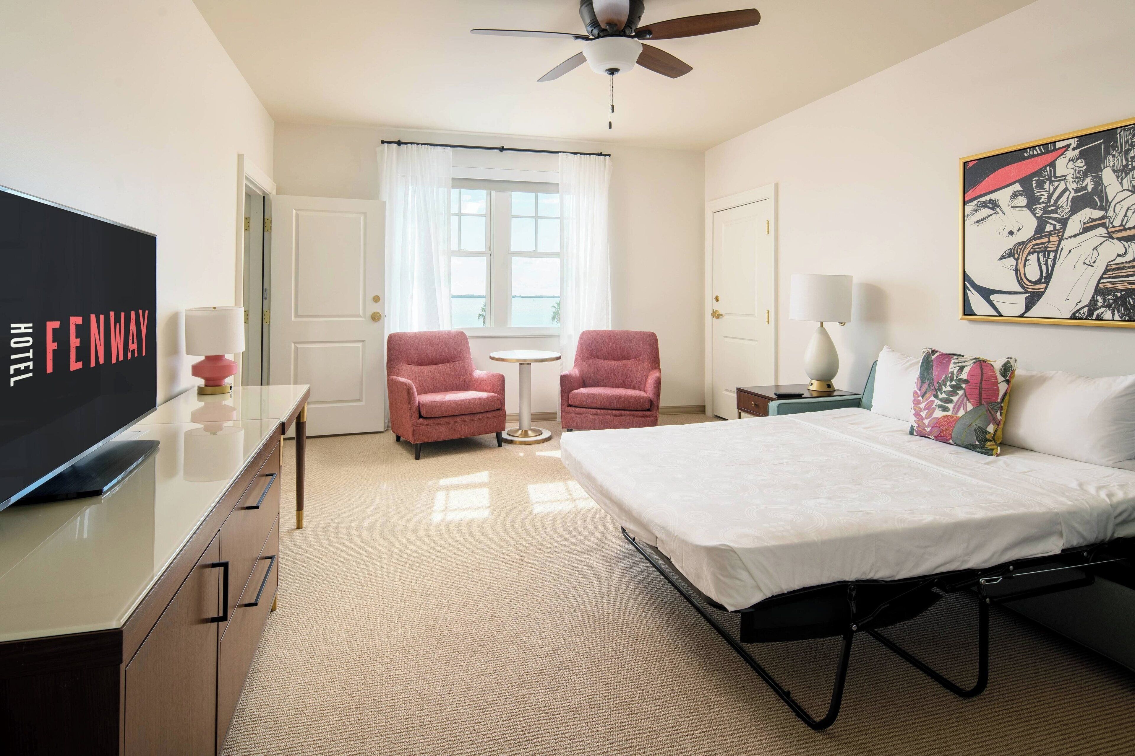 A hotel room with a bed, two pink armchairs, a small table, a TV, a ceiling fan, and a window with curtains.