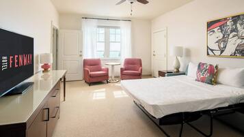 A hotel room with a bed, two pink armchairs, a small table, a TV, a ceiling fan, and a window with curtains.