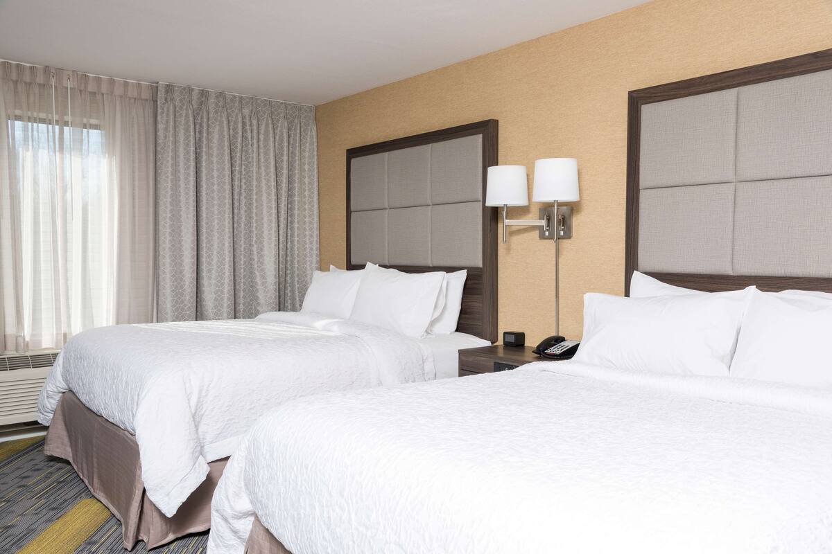A hotel room with two beds, a large window with curtains, a wall-mounted lamp, and a telephone on the nightstand.