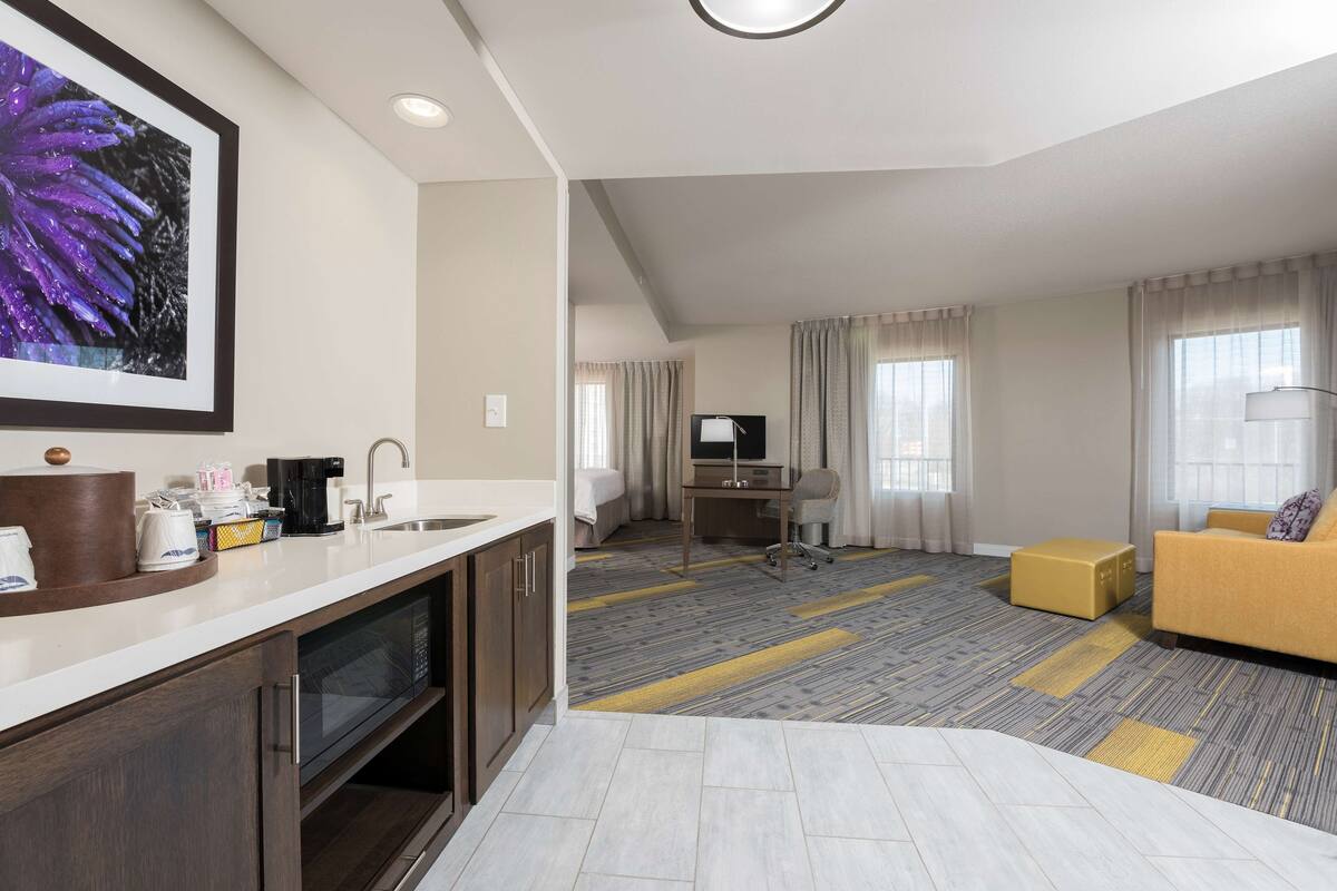 A hotel room with a kitchenette, a living area with a sofa and a TV, and a large window with curtains.