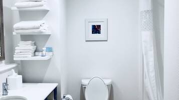 Une salle de bain moderne avec un lavabo, des toilettes et une douche, tous de couleur blanche. On y trouve des étagères avec des serviettes et des articles de toilette.