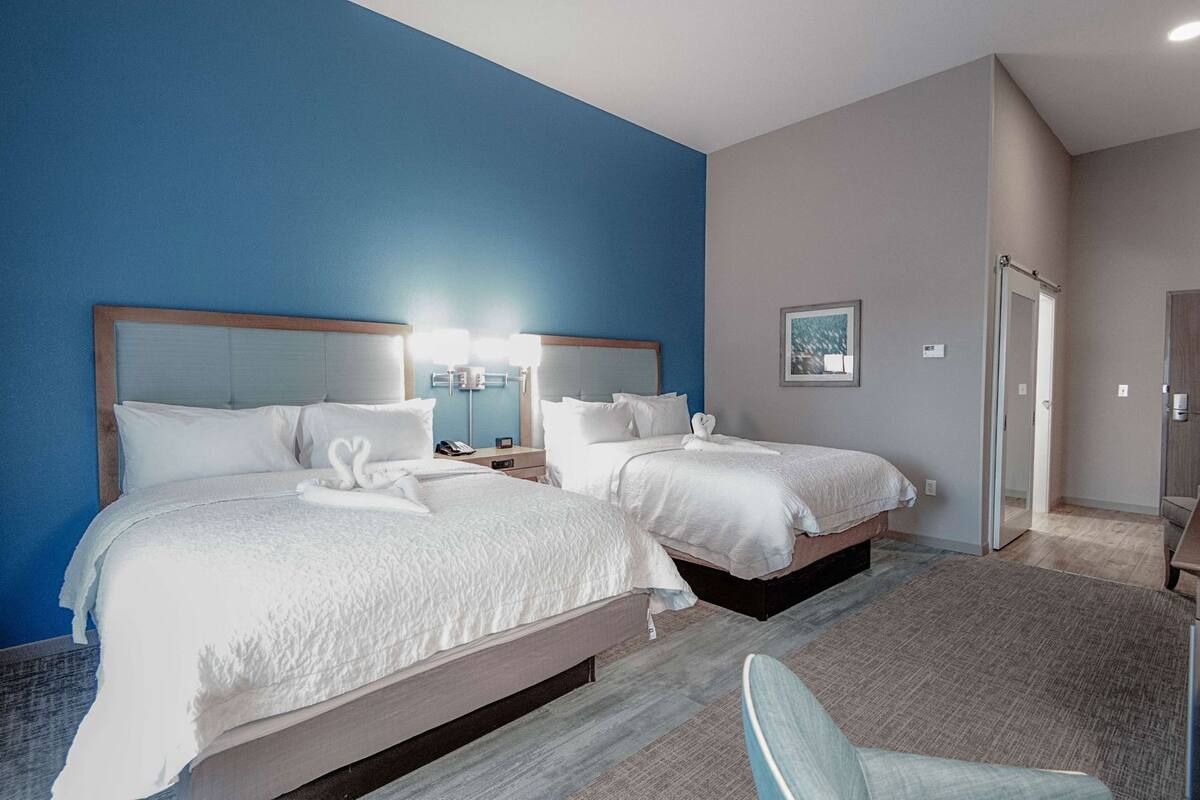 A hotel room with two beds, a nightstand with a lamp, a chair, and a door leading to another room.