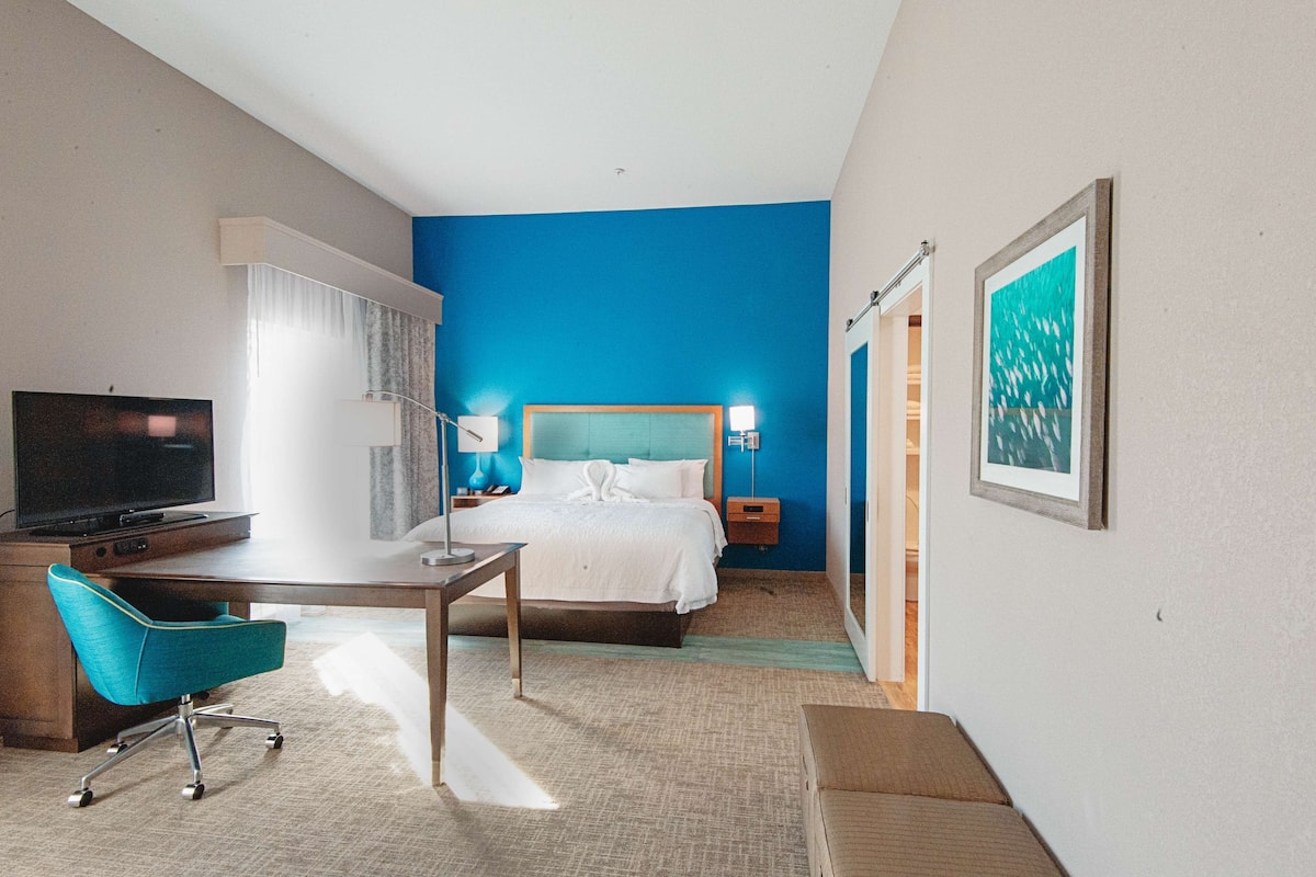 Una habitación de hotel con cama, escritorio, silla, televisor y una obra de arte en la pared.