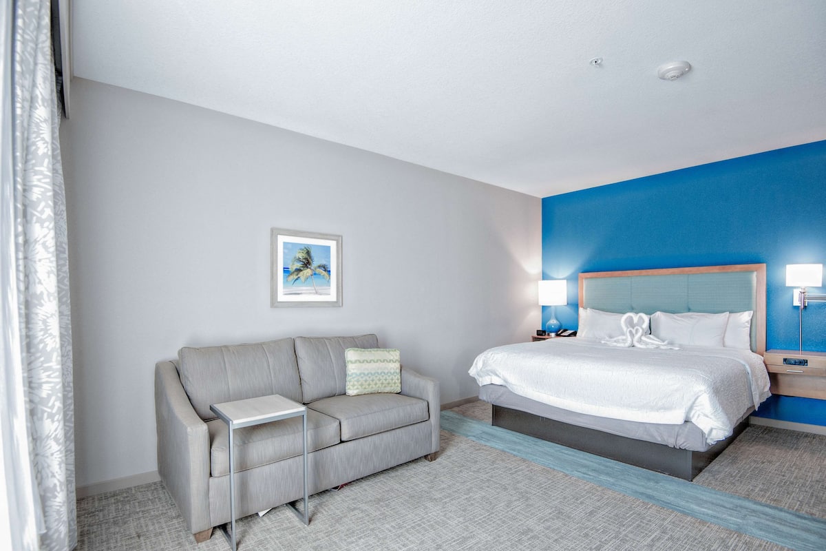 Una habitación de hotel con cama, sofá, mesita de noche y un cuadro colgado en la pared.