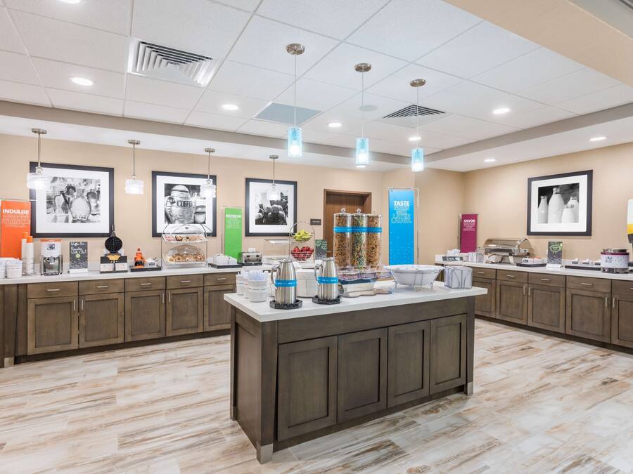 Un área de cocina moderna con una isla central, equipada con diversos electrodomésticos y alimentos. Hay varias luces colgantes y cuadros en las paredes.