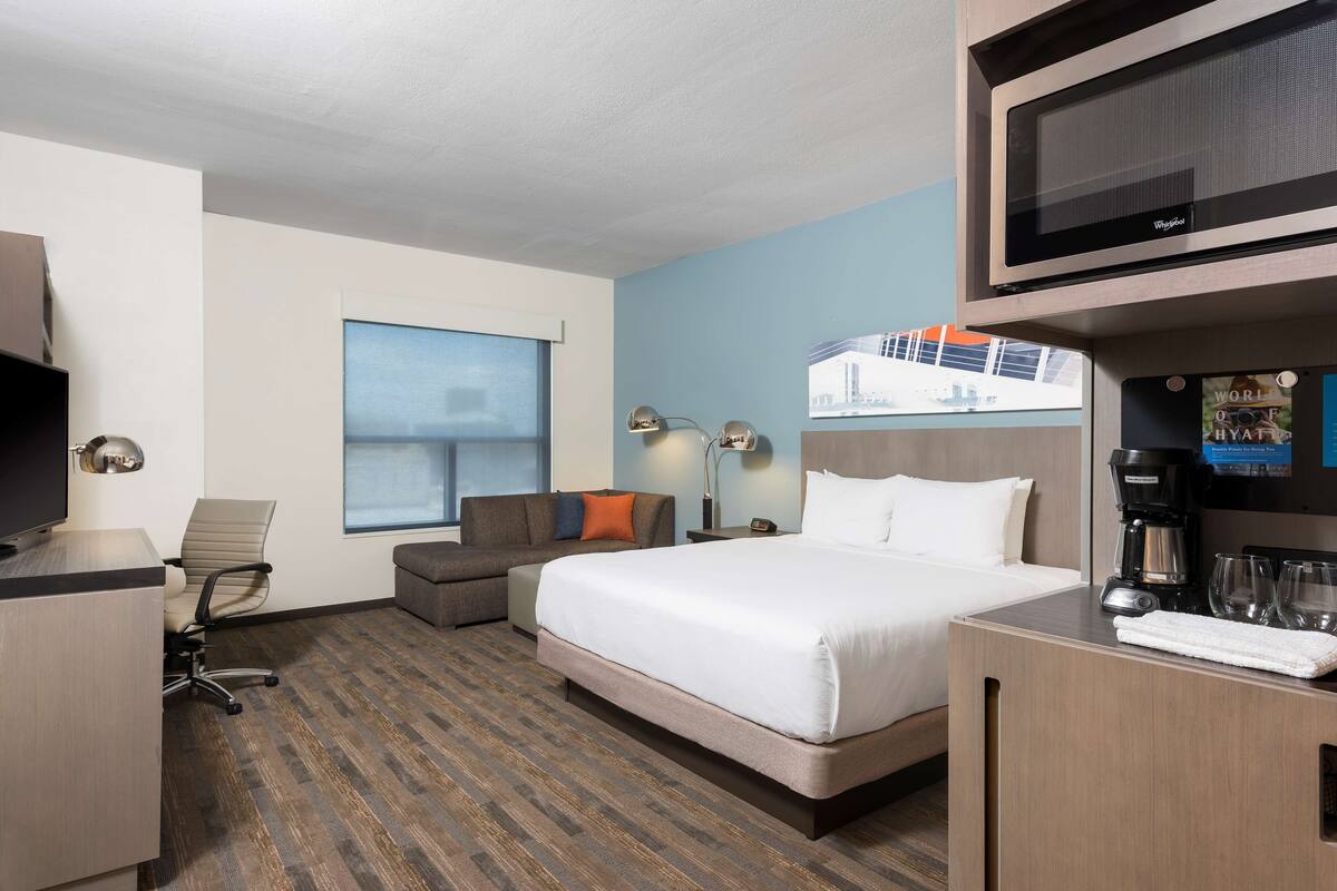 Habitación de hotel con cama, escritorio, silla, televisor, cafetera y ventana con vista a un edificio.