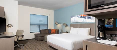 Quarto de hotel com cama, escrivaninha, cadeira, televisão, cafeteira e janela com vista para um edifício.