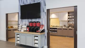 A modern kitchen with a flat-screen TV mounted above a coffee station, wooden cabinets, and a tiled backsplash.
