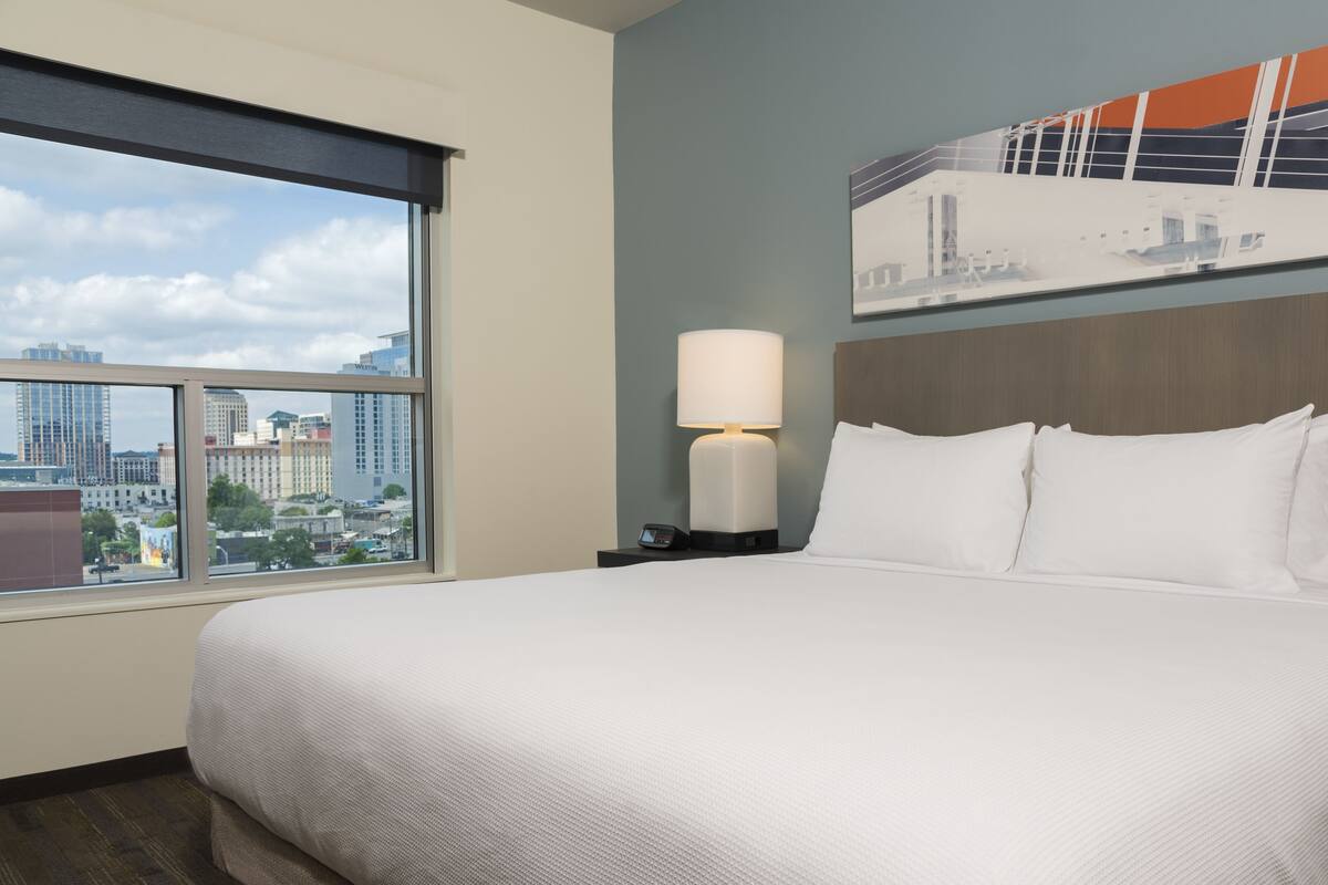 Habitación de hotel con una cama grande, una mesita de noche con lámpara, vistas a la ciudad a través de una ventana y un cuadro en la pared.