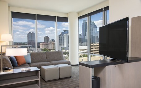 A modern hotel room with a large window, a flat-screen TV, a sofa, a coffee table, and a lamp.