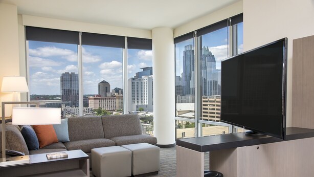 A modern hotel room with a large window, a flat-screen TV, a sofa, a coffee table, and a lamp. - Hyatt House Austin/Downtown (Austin)