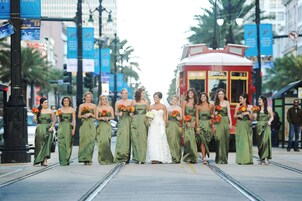 A group of people in green dresses and a white wedding dress standing on a street with a tram in the background.