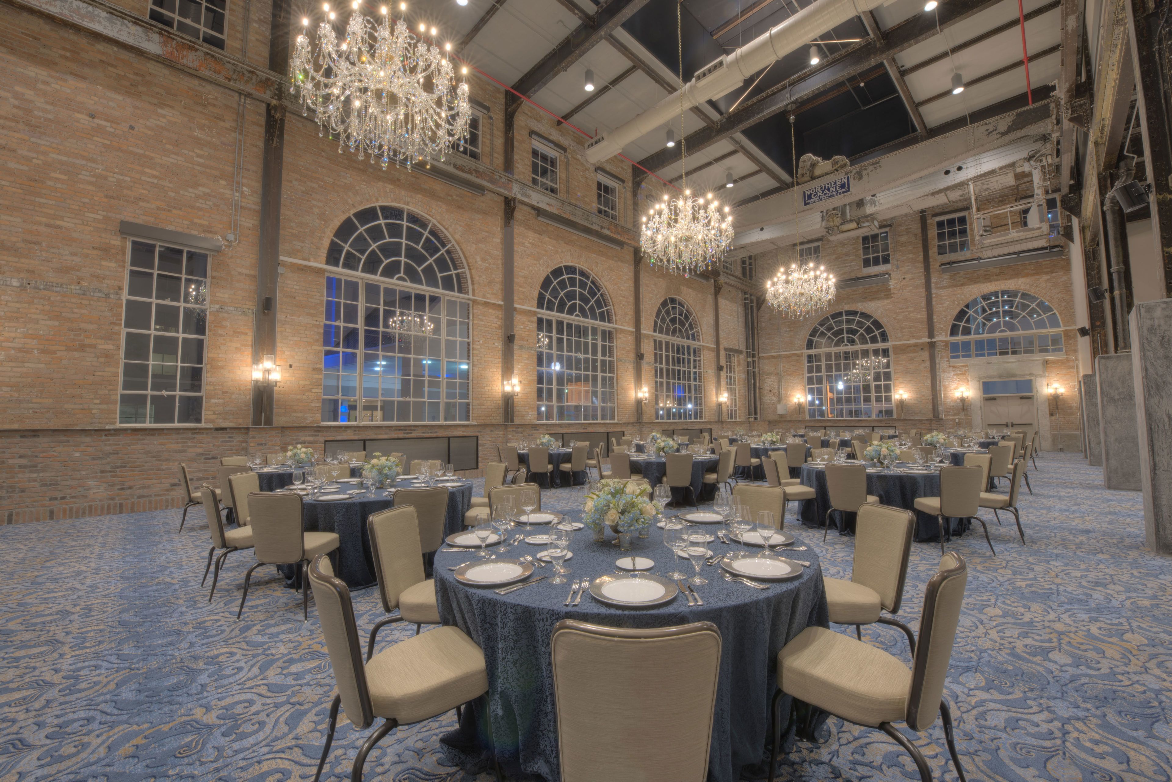 Un salón de banquetes con mesas redondas dispuestas para comer, amplias ventanas arqueadas y candelabros colgantes.