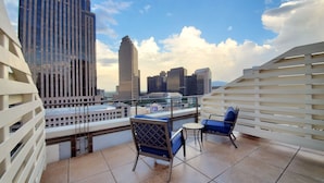 A rooftop terrace with a view of city skyscrapers, a white railing, and blue chairs.