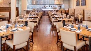 A spacious dining area with multiple tables and chairs, a visible kitchen in the background, and a brick wall on one side.