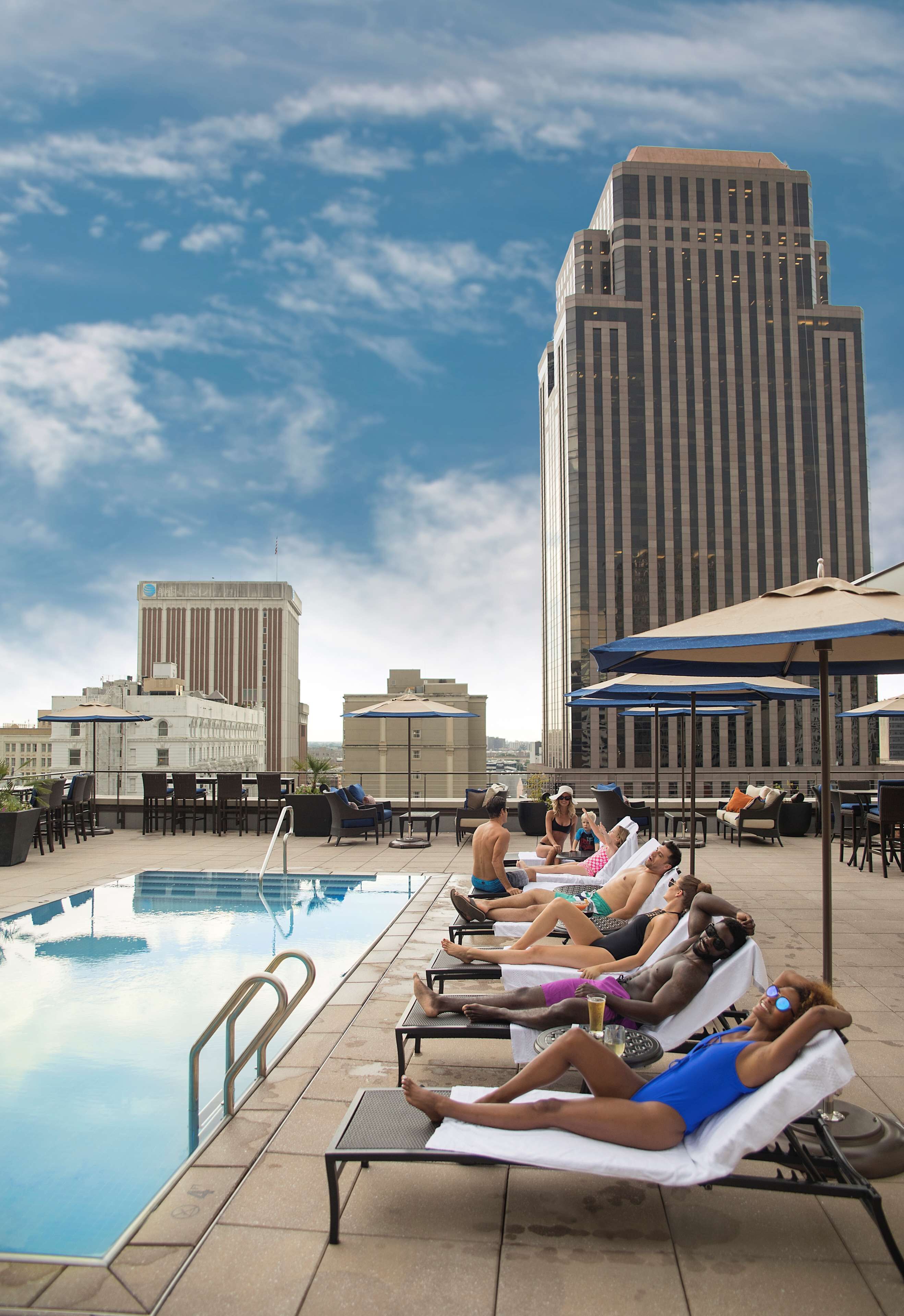 A rooftop pool area with lounge chairs and umbrellas, overlooking a cityscape with tall buildings.