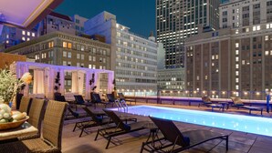 A rooftop pool area with lounge chairs and a dining table, surrounded by high-rise buildings.