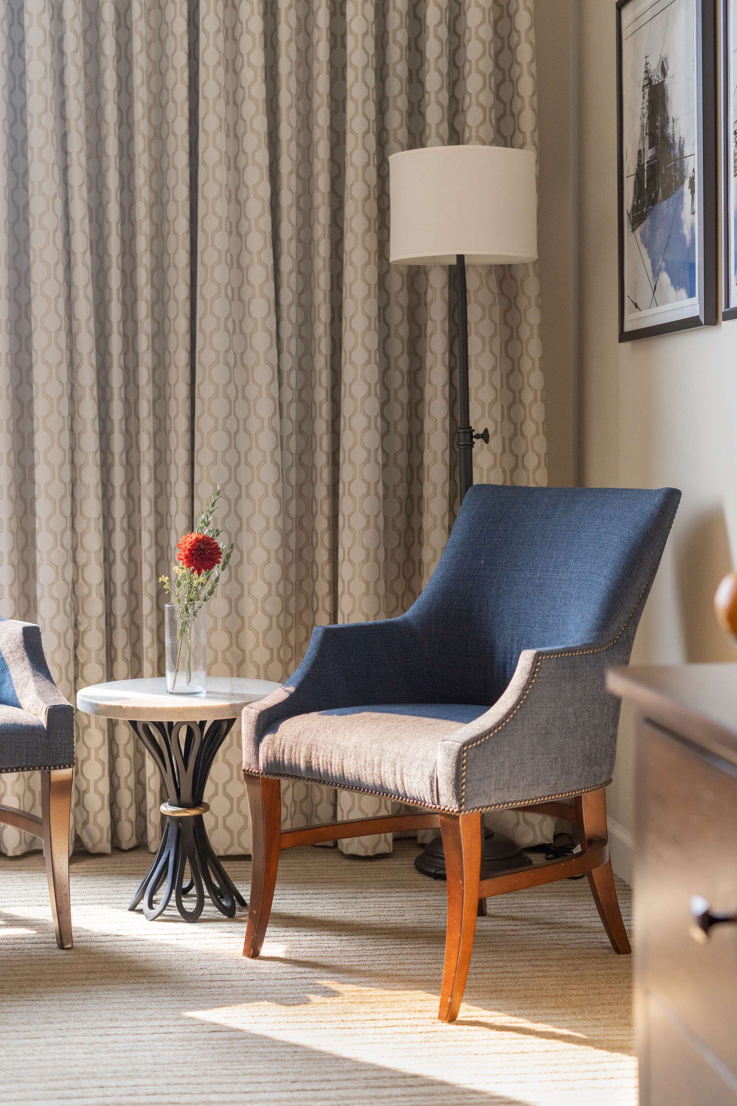 Una habitación con un sillón azul, una mesa redonda con un florero y una lámpara, además de una cortina estampada.