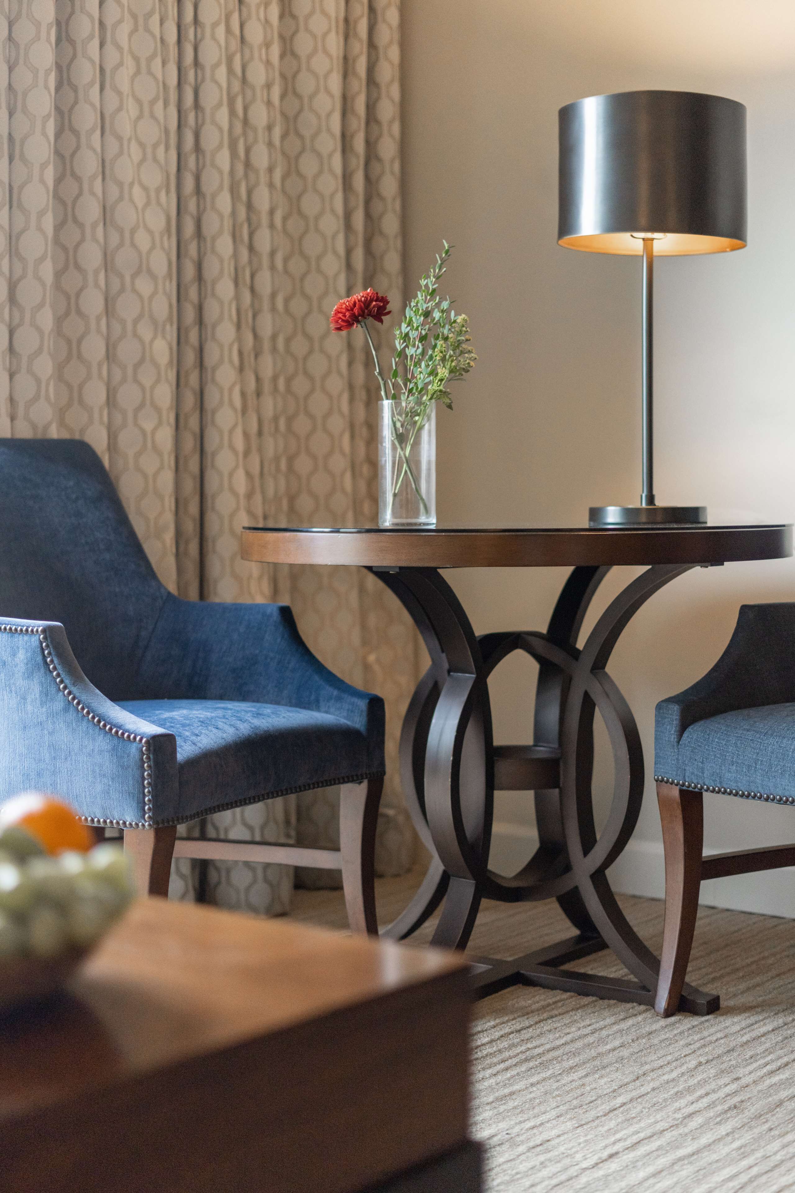 A hotel room with a round table, two blue armchairs, a vase with flowers, and a lamp.