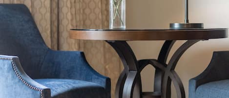 A hotel room with a round table, two blue armchairs, a vase with flowers, and a lamp.