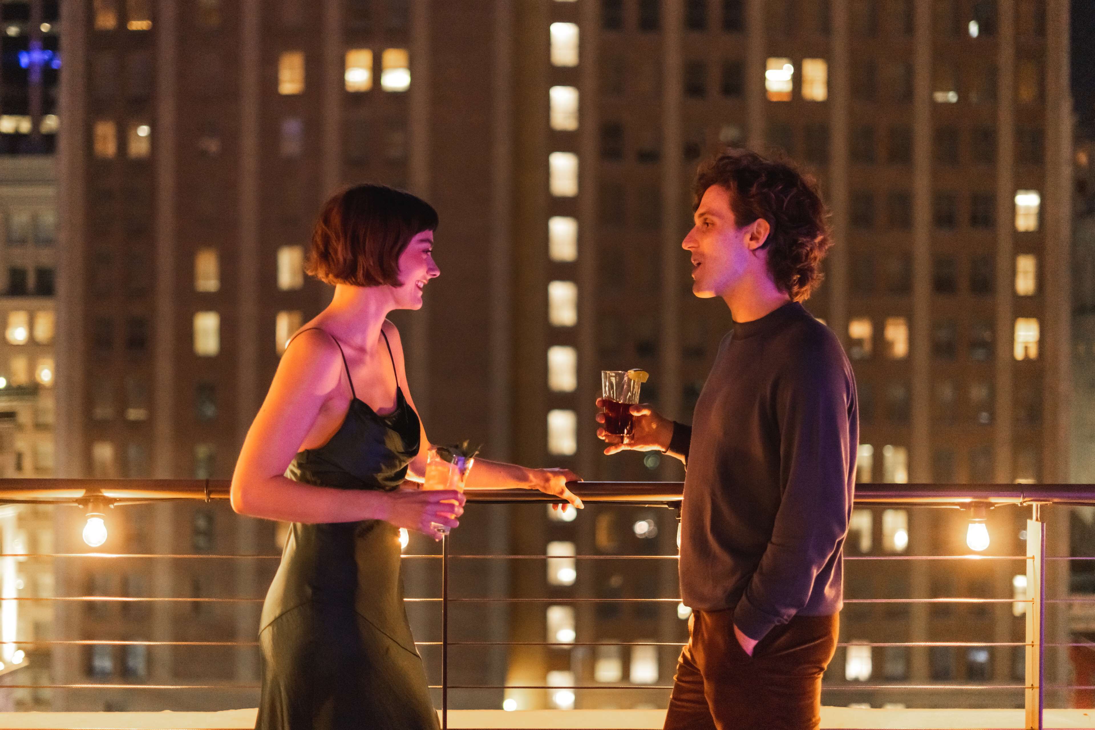 Two people holding drinks on a balcony with a cityscape in the background.