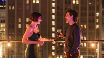 Two people holding drinks on a balcony with a cityscape in the background.