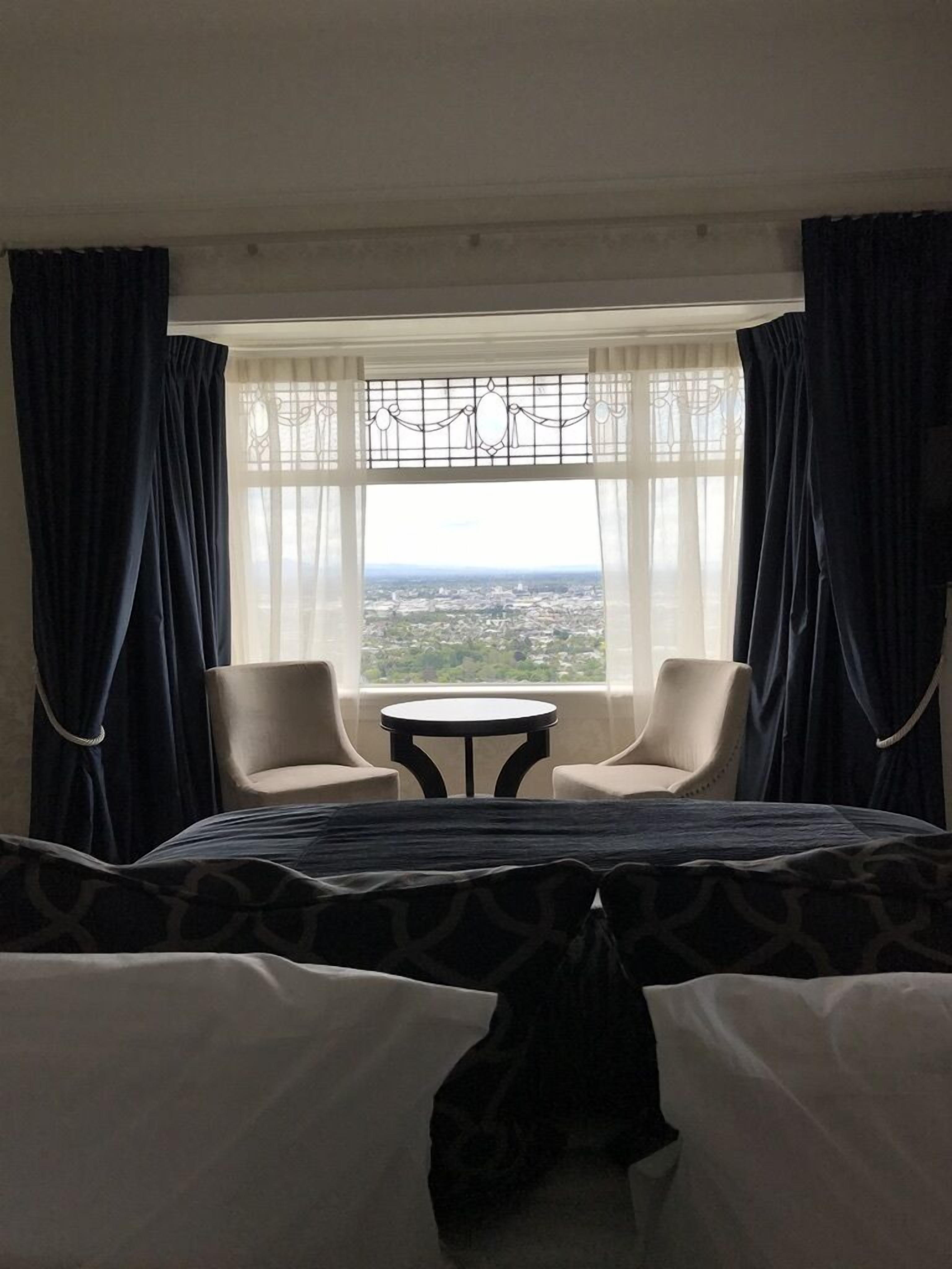 A hotel room with a bed, two chairs, a small table, and a large window offering a city view.