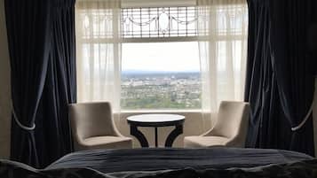 A hotel room with a bed, two chairs, a small table, and a large window offering a city view.