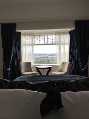 A hotel room with a bed, two chairs, a small table, and a large window offering a city view.