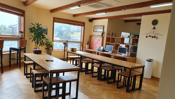 A spacious room with wooden tables and benches, large windows, and a bookshelf with various items. - dodo27 Guest House (Yeosu)