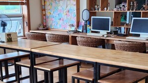 A spacious room with multiple wooden tables and benches, a ceiling fan, and a wall decorated with notes and pictures. - dodo27 Guest House (Yeosu)