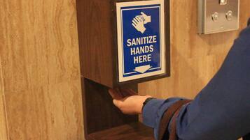 A hand sanitizer dispenser mounted on a wooden panel with a sign instructing to sanitize hands.