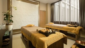 Habitación de hotel con dos camas, un ventanal con cortinas, una planta en maceta y una bandeja con toallas y artículos de aseo.