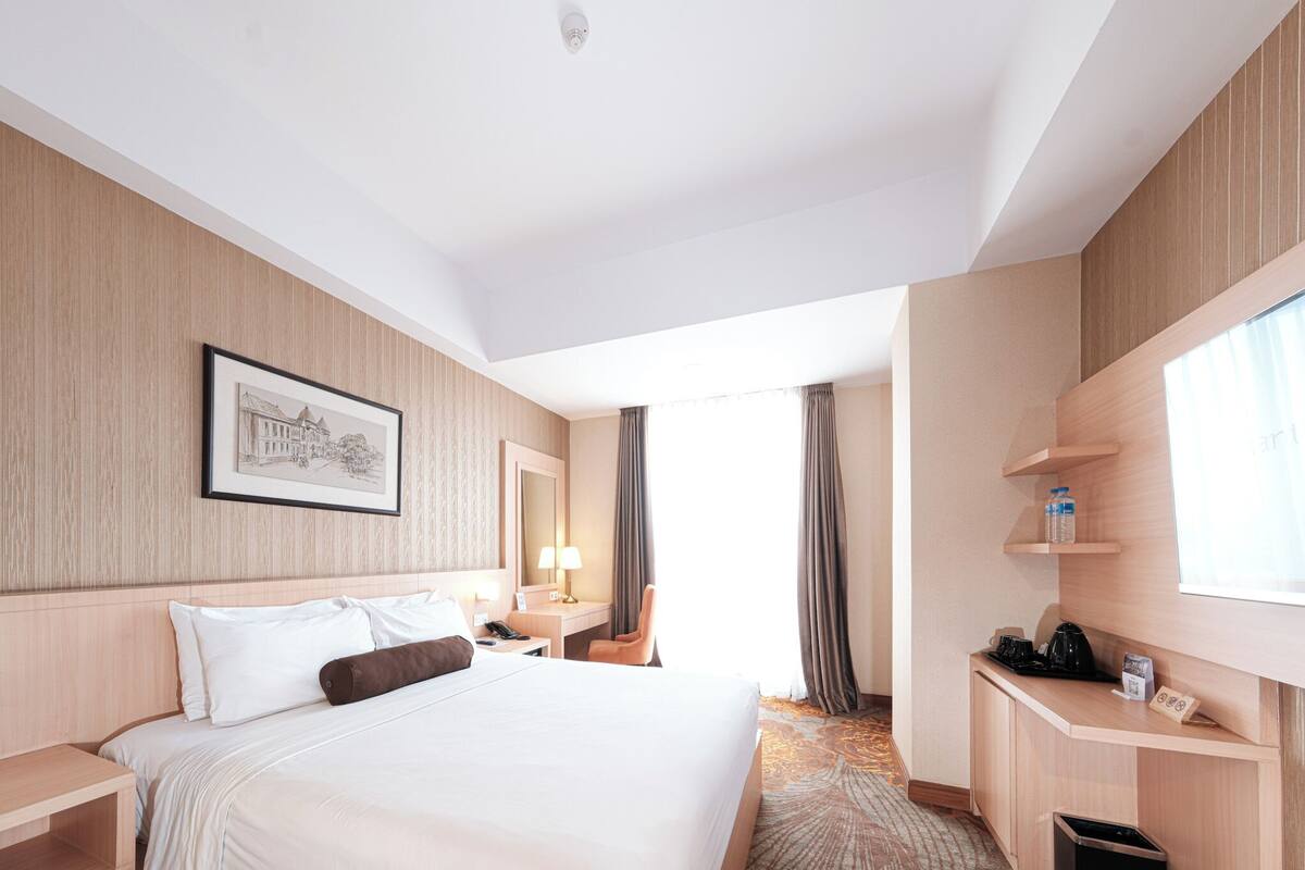 A hotel room with a large bed, a desk, a chair, a small table with a lamp, a picture on the wall, and a window with curtains.