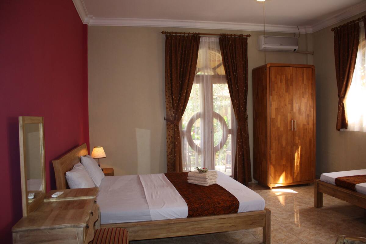 A hotel room with a bed, a wooden wardrobe, a nightstand, a mirror, and a window with curtains.