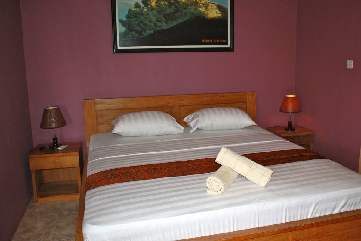 A neatly made bed with white linens and a rolled towel, two pillows, wooden headboard, two bedside tables with lamps, and a framed picture of a mountain on the wall.