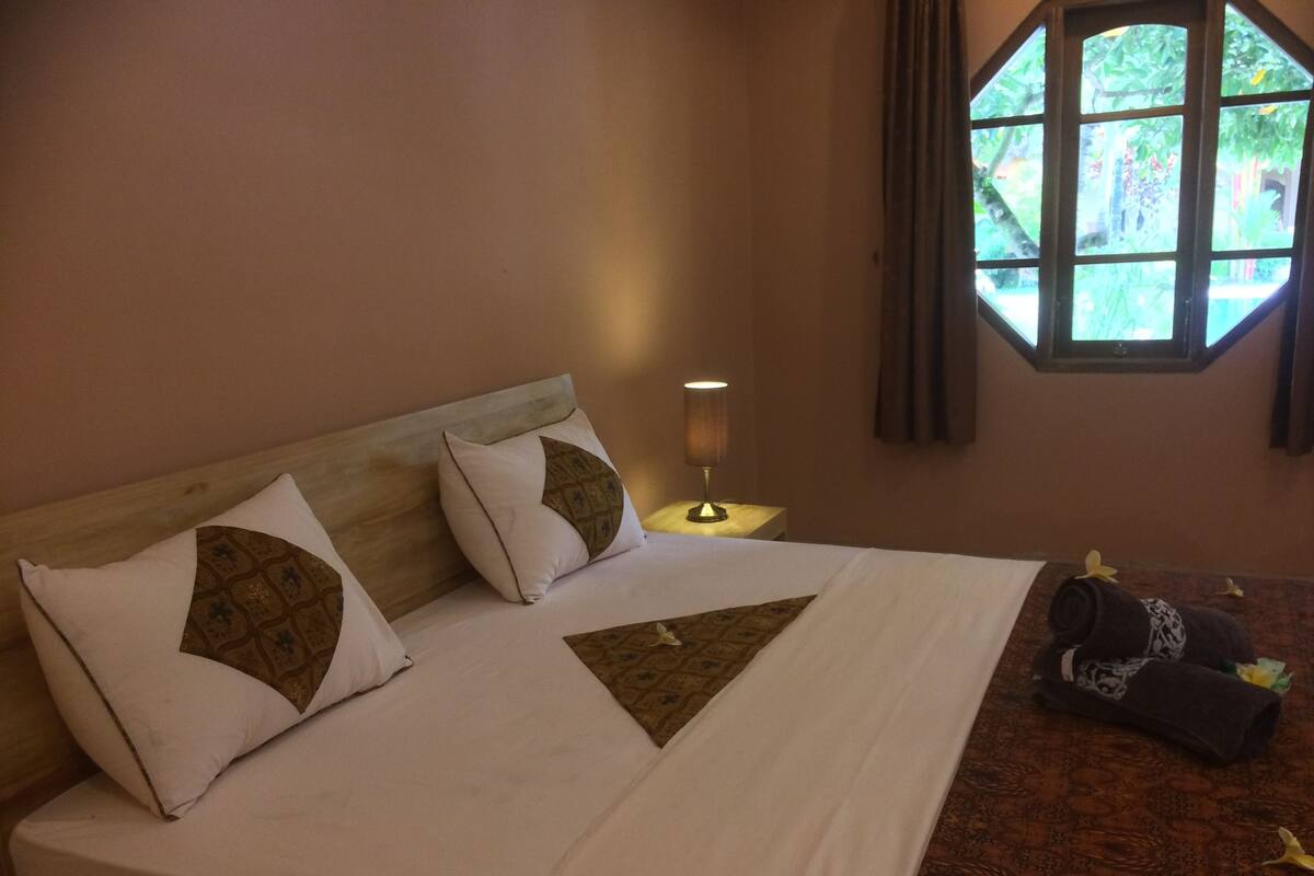 A hotel room with a bed, pillows, a nightstand with a lamp, a window with curtains, and a patterned carpet.